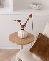 The Artificial Branch Berries 64 cm pink - en hvid vase fyldt med lyserøde kunstige bærgrene og grønne blade - står på et lille rundt bord ved siden af en beige stol med en brun pude i et hyggeligt, minimalistisk rum.