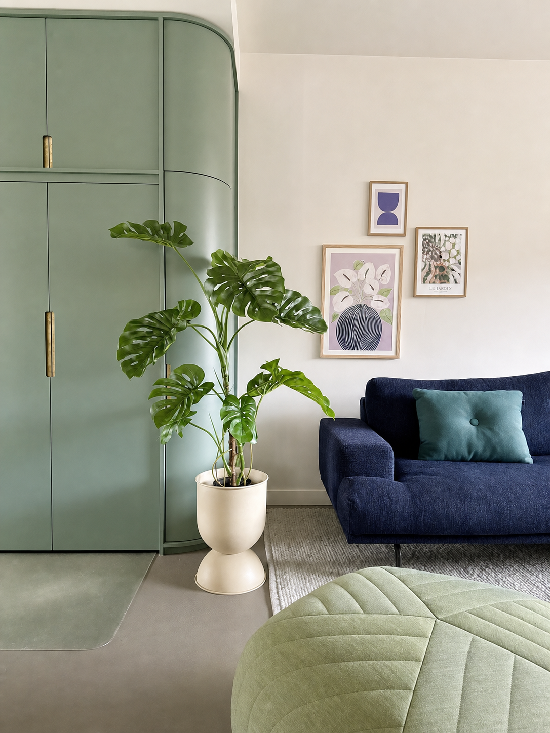 Een moderne woonkamer met een groene inbouwkast, Kunstplant Monstera 120 cm, een blauwe bank met een groenblauw kussen, abstracte kunst aan de muur en een groene poef op een licht vloerkleed.