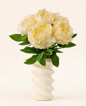 En bølget, struktureret hvid vase indeholder Silk kunstig blomsterbuket Peony White - store cremefarvede kunstige pæonblomster med grønne blade mod en almindelig lys baggrund.