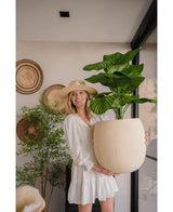 En kvinde i hvid kjole og hat smiler, mens hun holder den kunstige Calathea UV 72 cm plante indendørs med vævede kurve og en anden plante i baggrunden.