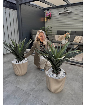 Een vrouw met lang blond haar knielt en glimlacht tussen twee Kunstplant Agava Pandora UV 85 cm potplanten op een betegelde patio, met buitenmeubilair en bloemen aan de muur op de achtergrond.