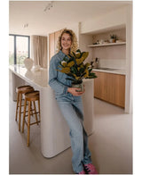En smilende person i denimtøj står i et moderne køkken med en kunstig magnolia-plante i en 46 cm krukke, en letplejende plante. Køkkenet, som er perfekt til indretning, byder på træskabe, en hvid køkkenø og to barstole.