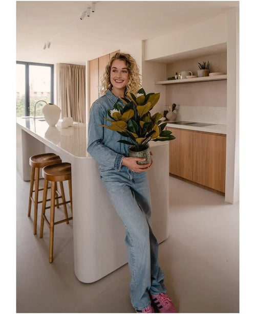 En smilende person i denimtøj står i et moderne køkken med en kunstig magnolia-plante i en 46 cm krukke, en letplejende plante. Køkkenet, som er perfekt til indretning, byder på træskabe, en hvid køkkenø og to barstole.