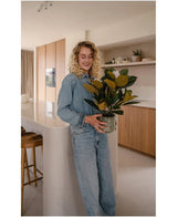 En kvinde med krøllet blond hår, klædt i denimtøj, står smilende i et moderne køkken med den kunstige magnolia-plante i en 46 cm krukke i hænderne, omgivet af træskabe og hylder – et ideelt valg for alle, der leder efter planter med lav vedligeholdelse for at forbedre deres stilfulde interiør.
