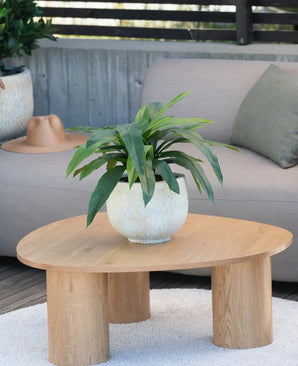 En kunstig plante Bird's Deluxe på 60 cm står på et rundt sofabord i træ foran en beige sofa, dekoreret med en grøn pude og en brun hat hængende over armlænet.