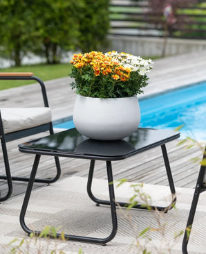 Et sort terrassebord med en Artificial Daisy UV 31 cm plante – en rund hvid potte med levende orange og hvide tusindfryd – står ved siden af ​​en swimmingpool, omgivet af havestole og med grønt i baggrunden.