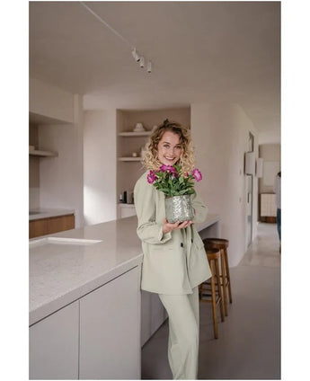 En kvinde i et lyst jakkesæt smiler, mens hun holder en kunstig blomst Petunia UV 25 cm mørk pink; hun står i et moderne køkken med neutrale toner og minimalistisk indretning.
