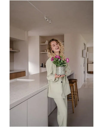 En smilende kvinde med krøllet hår, iført et lyst jakkesæt, står i et moderne, minimalistisk køkken med en Artificial Flower Petunia UV 25 cm mørk pink med levende lyserøde blomster.