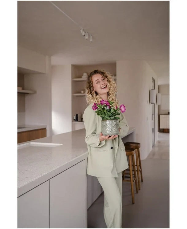 En smilende kvinde med krøllet hår, iført et lyst jakkesæt, står i et moderne, minimalistisk køkken med en Artificial Flower Petunia UV 25 cm mørk pink med levende lyserøde blomster.