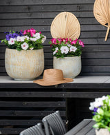 To store keramikkrukker med en kunstig blomst Petunia UV 25 cm mørk pink, med levende lyserøde blomster, står på et bord mod en mørk trævæg. Der står en stråhat på bordet og tørrede palmeblade hænger på væggen over blomsterne.
