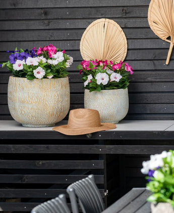 To store keramikkrukker med en kunstig blomst Petunia UV 25 cm mørk pink, med levende lyserøde blomster, står på et bord mod en mørk trævæg. Der står en stråhat på bordet og tørrede palmeblade hænger på væggen over blomsterne.