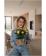 En kvinde med krøllet blond hår, klædt i denimtøj, sidder i et moderne køkken med Artificial Plant Mini Rose UV 35 cm gul i hænderne og kigger direkte ind i kameraet.