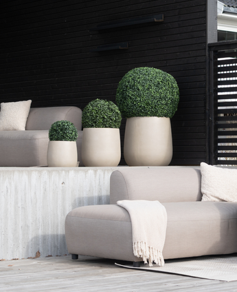 Een moderne patio heeft beige zitkussens, een crèmekleurige deken en vier Kunstplant Buxus romeinse stijl UV D30cm vormsnoei planten in lichtgekleurde potten tegen een muur met donker houten panelen.