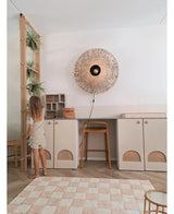En ung pige i lys kjole står ved siden af ​​en hylde, hvorpå den kunstige hængeplante 'Frosted Fern on ball 76 cm' og andre hængeplanter står, i et moderne rum med beige skabe, en høj skammel, en flettet væglampe og et ternet tæppe på gulvet.