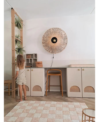 En ung pige i lys kjole står ved siden af ​​en hylde, hvorpå den kunstige hængeplante 'Frosted Fern on ball 76 cm' og andre hængeplanter står, i et moderne rum med beige skabe, en høj skammel, en flettet væglampe og et ternet tæppe på gulvet.