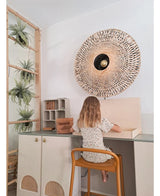 Et lille barn med skulderlangt hår sidder ved et skrivebord i et lyst rum, med ryggen vendt mod beskueren. Bag hende står en vævet væglampe, nogle skabe og den kunstige hængeplante 'Frosted Fern on ball 76 cm', puttet blandt planterne på en træstige, ved siden af ​​små hylder med pænt arrangerede ting.