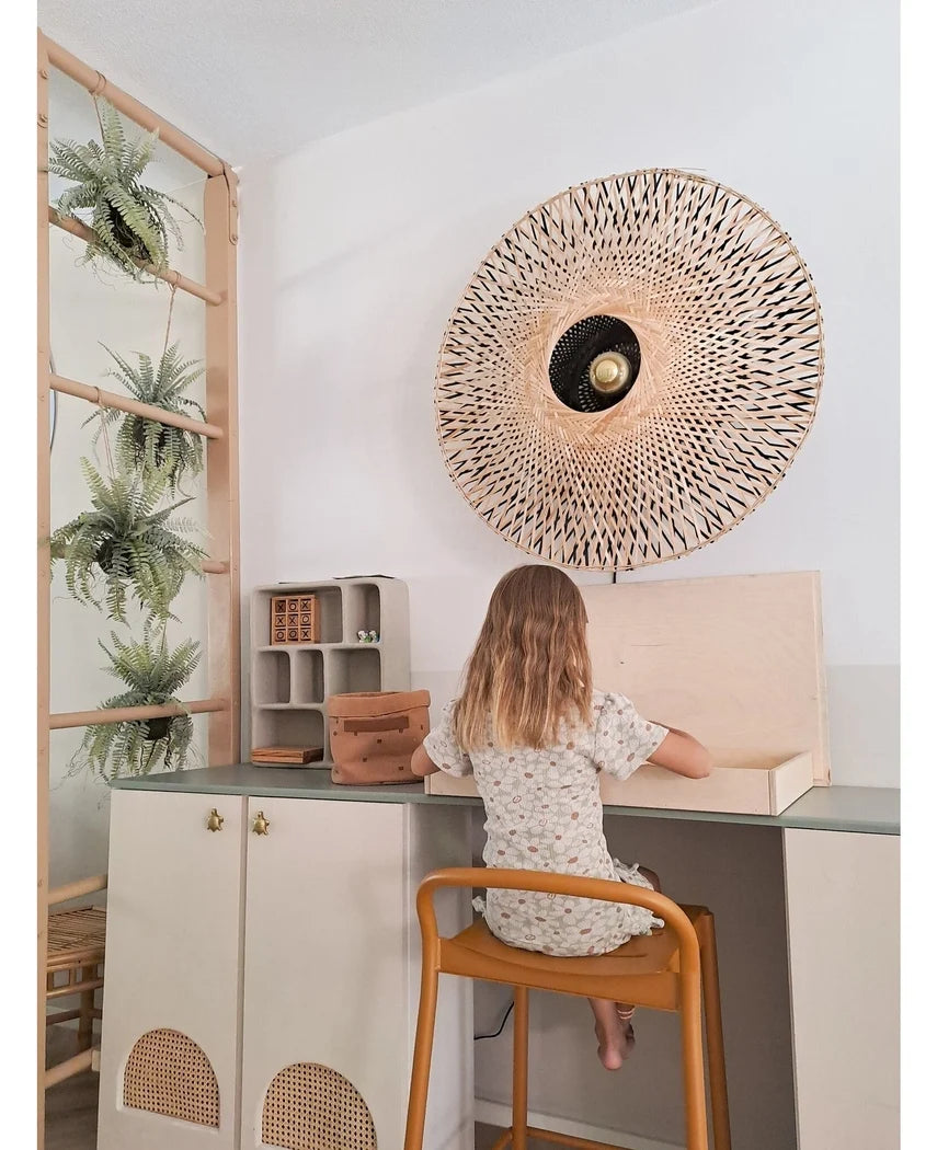 Et lille barn med skulderlangt hår sidder ved et skrivebord i et lyst rum, med ryggen vendt mod beskueren. Bag hende står en vævet væglampe, nogle skabe og den kunstige hængeplante 'Frosted Fern on ball 76 cm', puttet blandt planterne på en træstige, ved siden af ​​små hylder med pænt arrangerede ting.