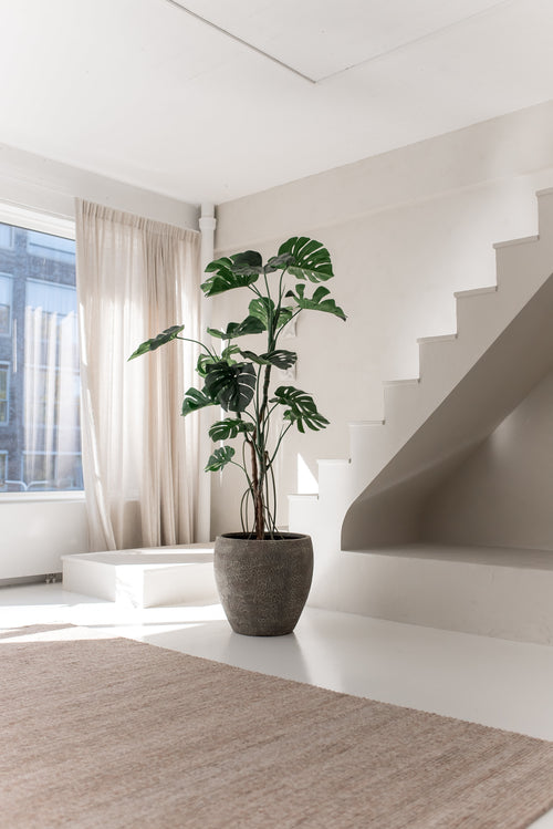 Den kunstige plante Monstera 210 cm står i en potte på et hvidt gulv ved siden af ​​en moderne trappe i et lyst, minimalistisk rum med naturligt lys, der kommer ind gennem beige gardiner.