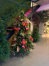 De Kunstkerstboom "Weideman" PE/PVC - Smart LED staat binnen bij een trap, versierd met roze en rode bloemen, gouden bladeren en roze ornamenten, omgeven door groen op een gepolijste betonnen vloer voor een stijlvolle kerstversiering.