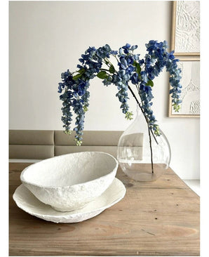 Een bloemstuk van Kunst Blauweregen van 115 cm staat in een doorzichtige vaas op een houten tafel, naast twee witte schalen met een structuur, terwijl de muur erachter versierd is met abstracte kunst.