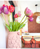 Een vaas met roze hartmotieven houdt Kunstbloem Franse Tulp 60 cm paars op een eettafel met stoelen met geweven rugleuning. Op de achtergrond dragen een ronde spiegel en een muur met bruin patroon bij aan het gezellige, moderne interieur.