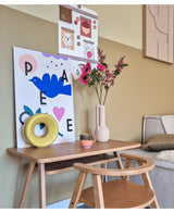 Een klein houten bureau met een stoel is versierd met Kunstbloem Catnip 75 cm roze, een mok, een gele ringsculptuur en kleurrijke kunstprints aan de muur erboven. Rechts is een bank gedeeltelijk zichtbaar.