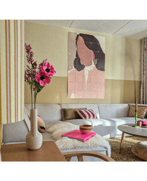 En moderne stue med en modulsofa, et lyserødt blomsterarrangement af Artificial Flower Catnip (75 cm) i en vase, en fluffy plaid og et abstrakt vægmaleri af en kvinde. Interiøret er præget af bløde farver og mønstrede accenter.