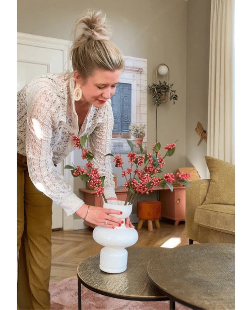 En kvinde med lyst hår i en knold arrangerer Kunsttak Berries 64 cm pink i en hvid vase på et rundt bord i en hyggelig stue med jordfarver og moderne indretning.