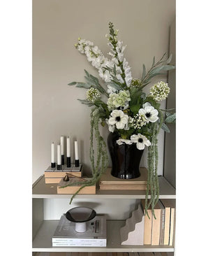 På en hylde mod en lys væg står en sort vase med Kunstblomsten Amaranthus 124 cm grøn, ved siden af ​​fem hvide lys, beige bøger og dekorative genstande i neutrale toner.