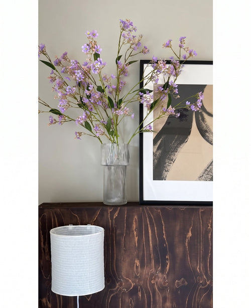 I en gennemsigtig vase er blomsterne i Kunsttak Daphne Spray 108 cm lilla placeret på en mørk træoverflade ved siden af ​​et moderne, abstrakt kunstværk i en ramme, med en hvid struktureret lampeskærm nedenunder.