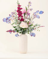 En hvid tekstureret vase viser "Silk Artificial Flowers Bouquet Amethyst Romance" med lyserøde pæoner, lilla og blå blomster, bordeauxrøde blomster og grønt løv mod en lys, neutral baggrund.
