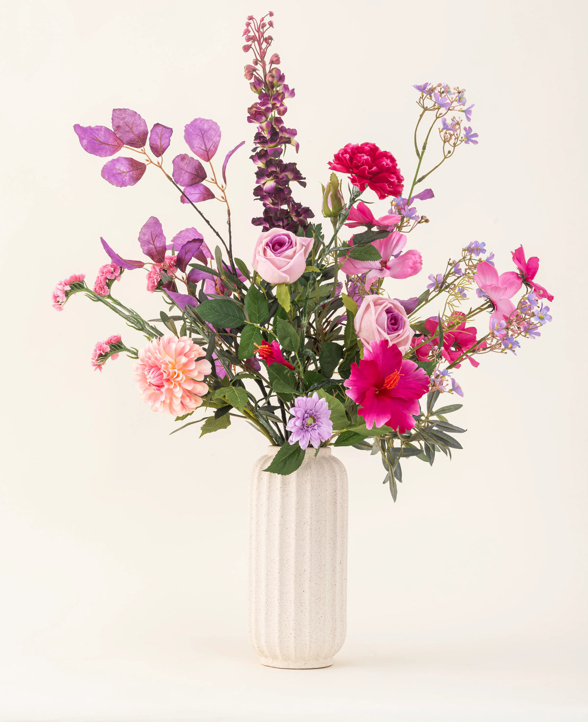 En hvid tekstureret vase indeholder Purple Dawn kunstige blomsterbuket i silke i pink, lilla og lavendel nuancer med grønt løv mod en lys, neutral baggrund.