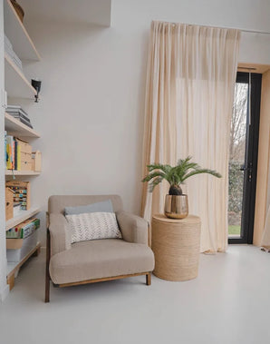 Et hyggeligt læsehjørne med en beige lænestol, et rundt sidebord i flet med kunstig Baby Cycas Palm 60 cm plante på, reoler og netgardiner, der hænger over en glasdør, der åbner ud til haven.