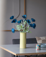 En 73 cm kunstig blomstervalmue i mørkeblå står i en gul vase på et bord, ledsaget af en lille, farverig stribet kop, i et moderne, blødt oplyst rum med minimalistisk indretning og neutrale toner.