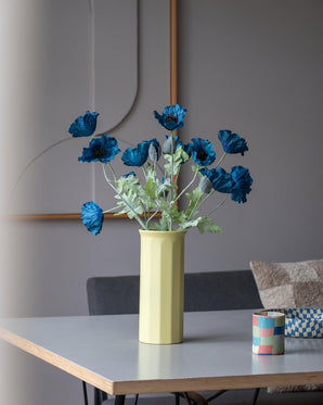 En 73 cm kunstig blomstervalmue i mørkeblå står i en gul vase på et bord, ledsaget af en lille, farverig stribet kop, i et moderne, blødt oplyst rum med minimalistisk indretning og neutrale toner.