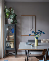 Den moderne spiseplads har et gråt vitrineskab, hvor service og planter er udstillet, minimalistisk vægdekoration og et bord, hvorpå Kunstig Blomstervalmue 73 cm mørkeblå står i en gul vase. Rummet fuldendes af en eklektisk blanding af stole, alt sammen mod en baggrund af grå vægge og et trægulv.
