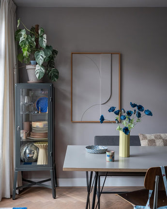 Den moderne spiseplads har et gråt vitrineskab, hvor service og planter er udstillet, minimalistisk vægdekoration og et bord, hvorpå Kunstig Blomstervalmue 73 cm mørkeblå står i en gul vase. Rummet fuldendes af en eklektisk blanding af stole, alt sammen mod en baggrund af grå vægge og et trægulv.