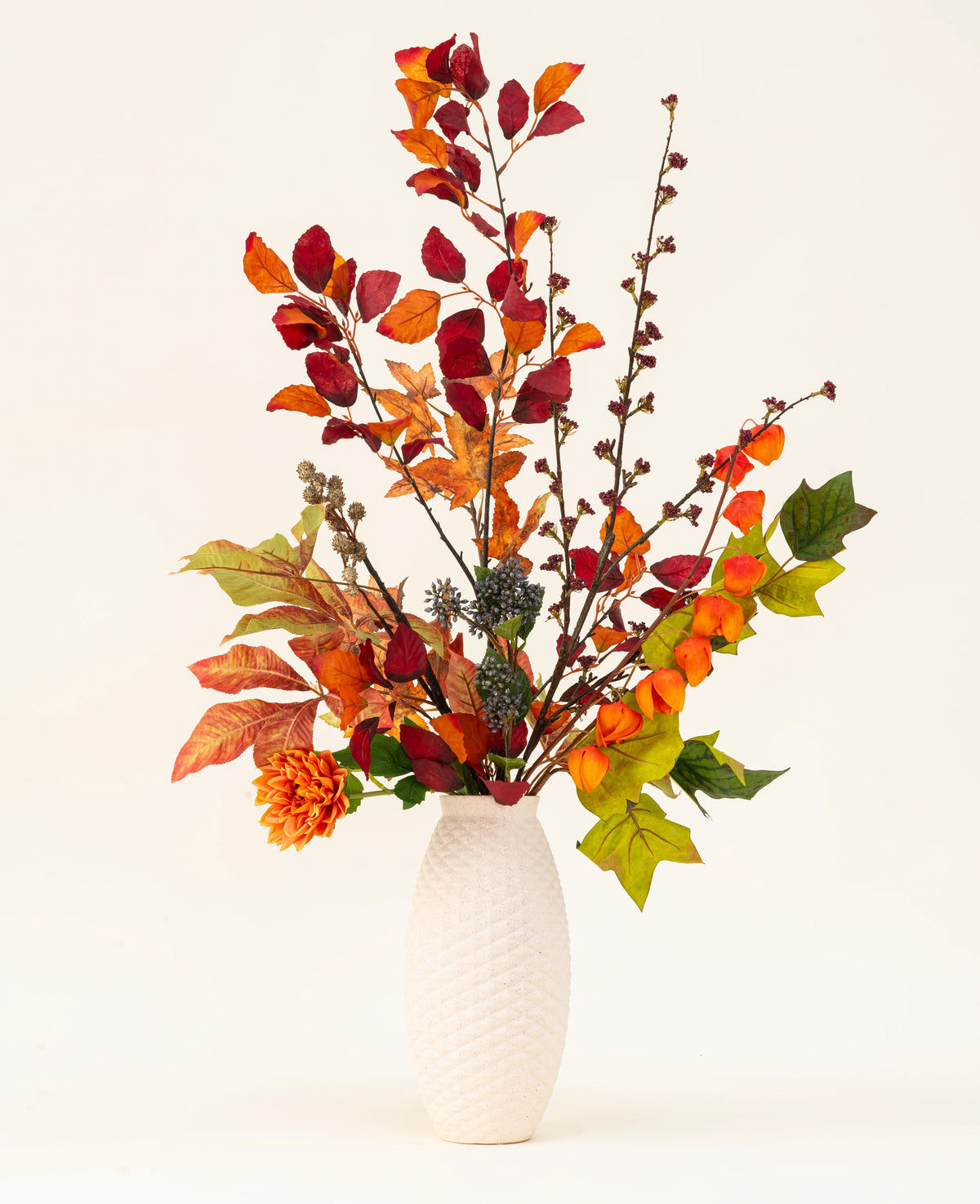En hvid tekstureret vase fremviser Chestnut Charm Artificial Flower Bouquet, med efterårsblade, orange blomster og grønt løv - et elegant kunstigt arrangement mod en almindelig lys baggrund.