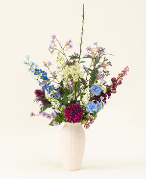 Een witte vaas met het Zijden kunstbloemen boeket Purple Rain toont levendige paarse en witte kunstbloemen en groen blad, een toonbeeld van Nederlands vakmanschap tegen een lichte achtergrond.