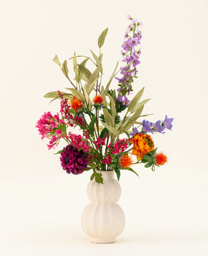 En hvid, tekstureret vase fremviser den farverige Svenski kunstige blomsterbuket i silke, med levende lilla, orange, pink og blå kunstige blomster med grønne blade, der lyser ethvert rum op mod en enkel lys baggrund.