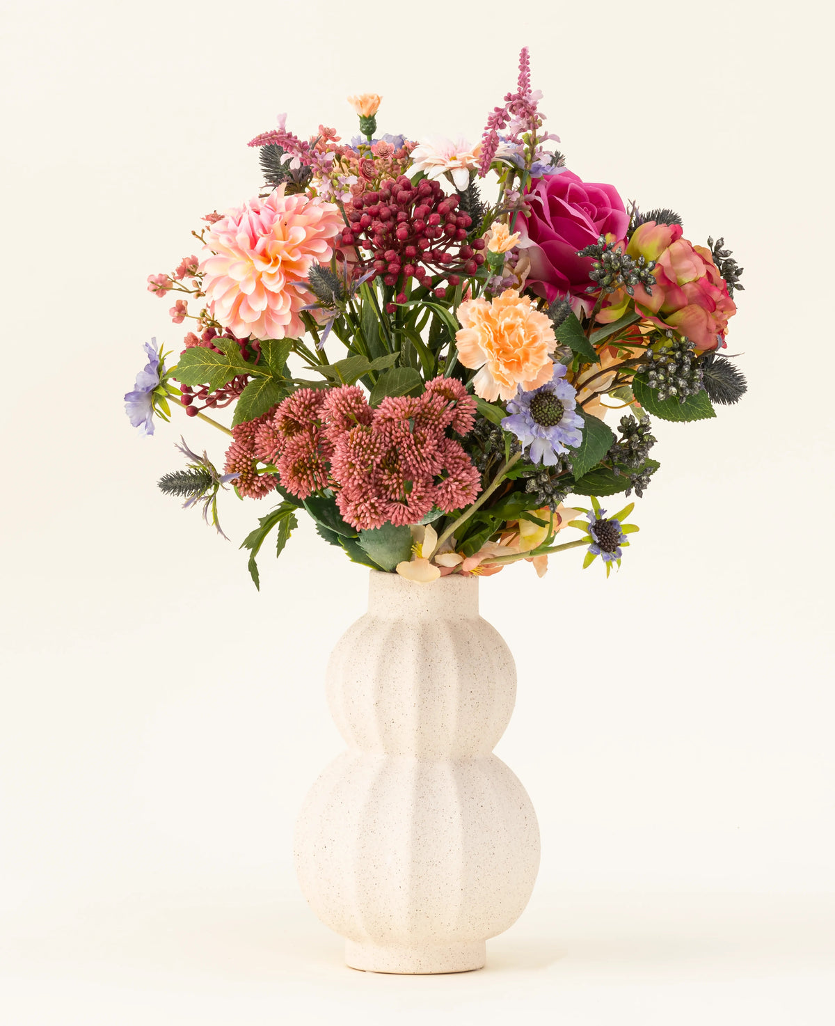 I en hvid, buet keramikvase er Pretty Pastel kunstige blomsterbuket i silke - en farverig blanding af bæredygtige blomster såsom lyserøde roser, georginer, lilla og orange blomster og grønt - smukt arrangeret mod en lys baggrund.