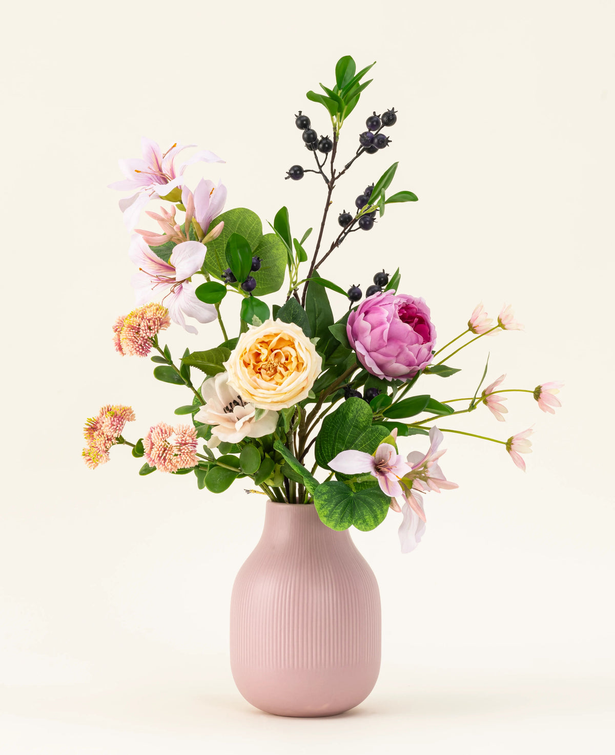 The Touch of Cloudlight kunstig silkeblomstbuket, præsenteret i en pink keramisk vase, indeholder lyserøde pæoner, gule roser, små blomster, grønt og grene med mørke bær på en lys baggrund.