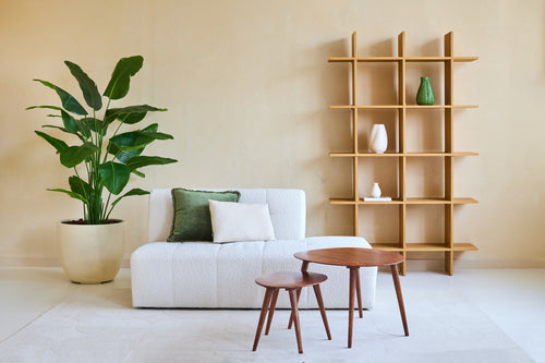 Minimalistisk stue med hvid sofa, to hynder, træborde og en stor kunstplante Strelitzia Real Touch Deluxe 180 cm for en tropisk atmosfære. En moderne åben bogreol i træ med vaser står op ad den beige væg.