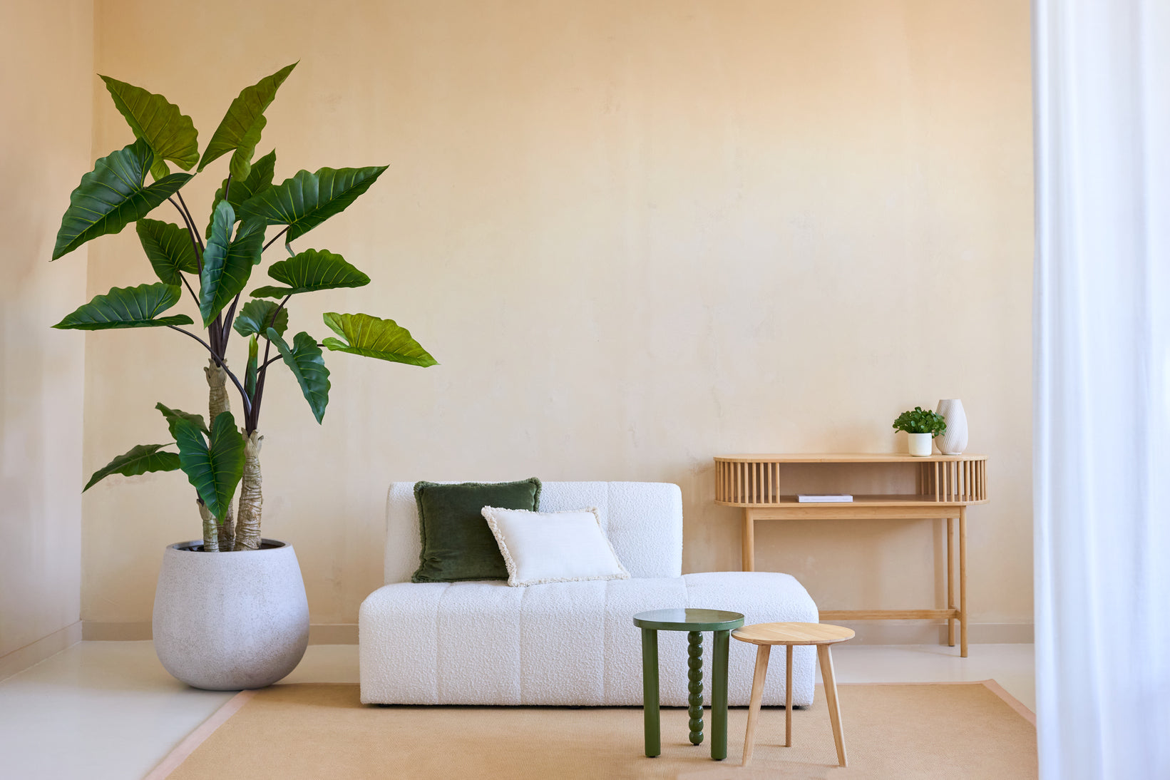 Een minimalistische woonkamer met de Kunstplant Calocasia 210 cm, een bank met witte textuur en groene en witte kussens, twee kleine bijzettafeltjes, een beige vloerkleed en een houten consoletafel tegen een lichtbeige muur.