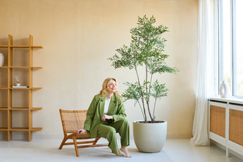 En kvinde i grønt jakkesæt smiler, mens hun sidder på en træstol ved siden af ​​Artificial Ruscus 180 cm planten i et lyst, moderne rum med beige vægge, store vinduer og minimalistisk indretning.