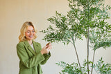 En smilende kvinde i en grøn blazer står indendørs og rører blidt ved bladene på en Artificial Ruscus 150 cm plante med tynde grene og frodigt grønt løv, der stikker ud mod en almindelig, lys baggrund.