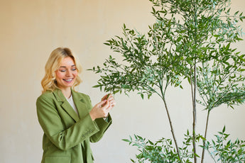 En smilende kvinde i en grøn blazer står indendørs og rører blidt ved bladene på en Artificial Ruscus 150 cm plante med tynde grene og frodigt grønt løv, der stikker ud mod en almindelig, lys baggrund.