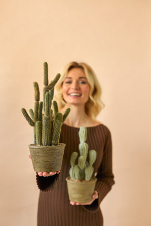En smilende kvinde i en brun kjole holder to Artificial Cactus Cereus 35 cm potteplanter, en stor og en lille, mod en almindelig beige baggrund.