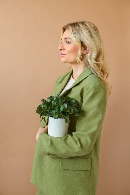 En kvinde med langt blondt hår i en lysegrøn blazer holder "Kunstig sukkulentplante 22 cm i dekorativ potte" mod sit bryst og står sidelæns foran en almindelig beige baggrund.