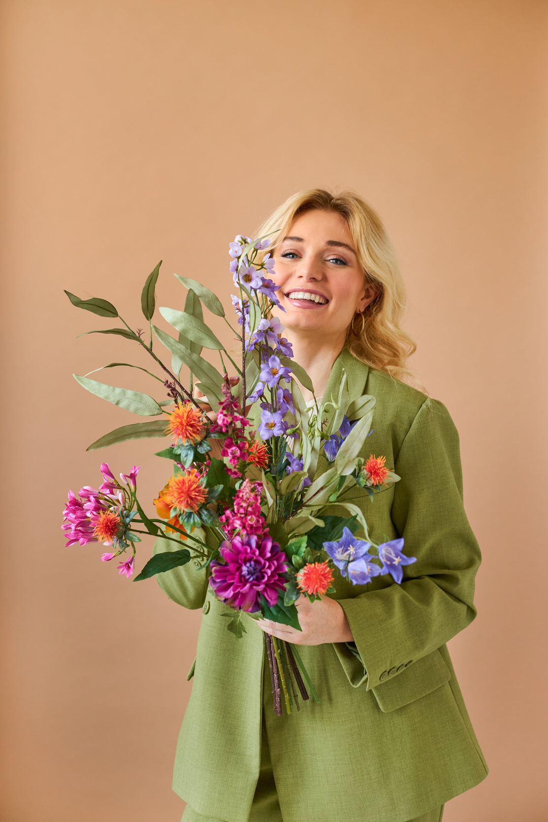 En smilende blond person i et grønt jakkesæt holder den livlige farverige Svenski kunstige blomsterbuket i silke mod en almindelig, lysebrun baggrund.